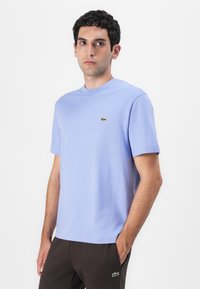 Helles blaues T-Shirt mit kurzen Ärmeln aus Baumwolle, das ein kleines grünes Logo auf der Brust trägt. Glatte Textur und klassisches Rundhals-Design.