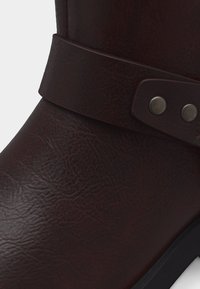 Καφέ σκούρο δερμάτινο μποτάκι μέχρι τον αστράγαλο με διακοσμητικό λουράκι και μεταλλικά καρφιά, που εμφανίζει υφή υλικού και μέρος της σόλας σε λευκό φόντο.