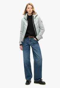 Chaqueta acolchada gris claro con capucha, suéter negro de punto, jeans de pierna ancha azules y zapatos negros; presenta una textura acolchada y detalles ajustables.