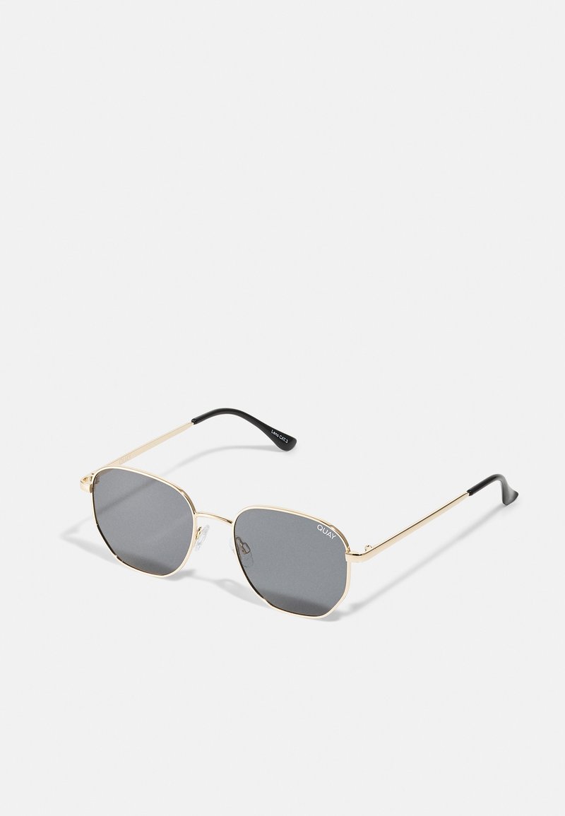 Gold metal frame sunglasses with dark gray lenses, angular square shape, black tips on the arms, and clear nose pads. Brand name visible on lens.