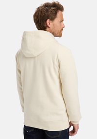 Sudadera de color crema, hecha de una tela suave, presenta una capucha con cordón y puños acanalados. Textura suave y corte relajado, detalles de costura visibles.