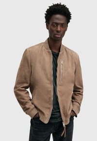 Giacca bomber in suede marrone con zip frontale, tasche laterali, polsini a coste e una texture liscia, indossata sopra una t-shirt nera.