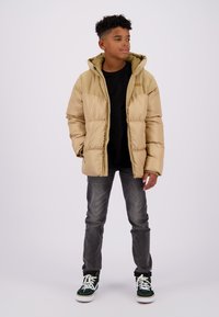 Beige pufferjack met capuchon, voorzien van een gewatteerd ontwerp. Gecombineerd met donkere jeans en groen/witte sneakers. Eenvoudige, functionele stijl.