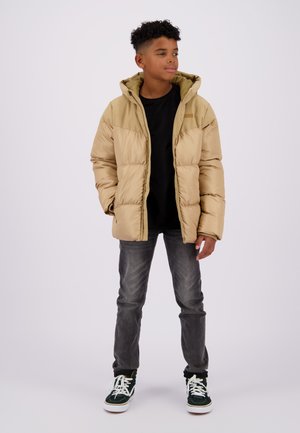 Beige pufferjack met capuchon, voorzien van een gewatteerd ontwerp. Gecombineerd met donkere jeans en groen/witte sneakers. Eenvoudige, functionele stijl.
