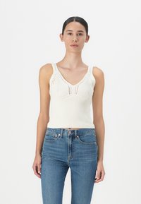 Hvit strikket tank topp med V-hals, ribbet tekstur og dekorativ åpenmønster. Kombinert med blå denim jeans.