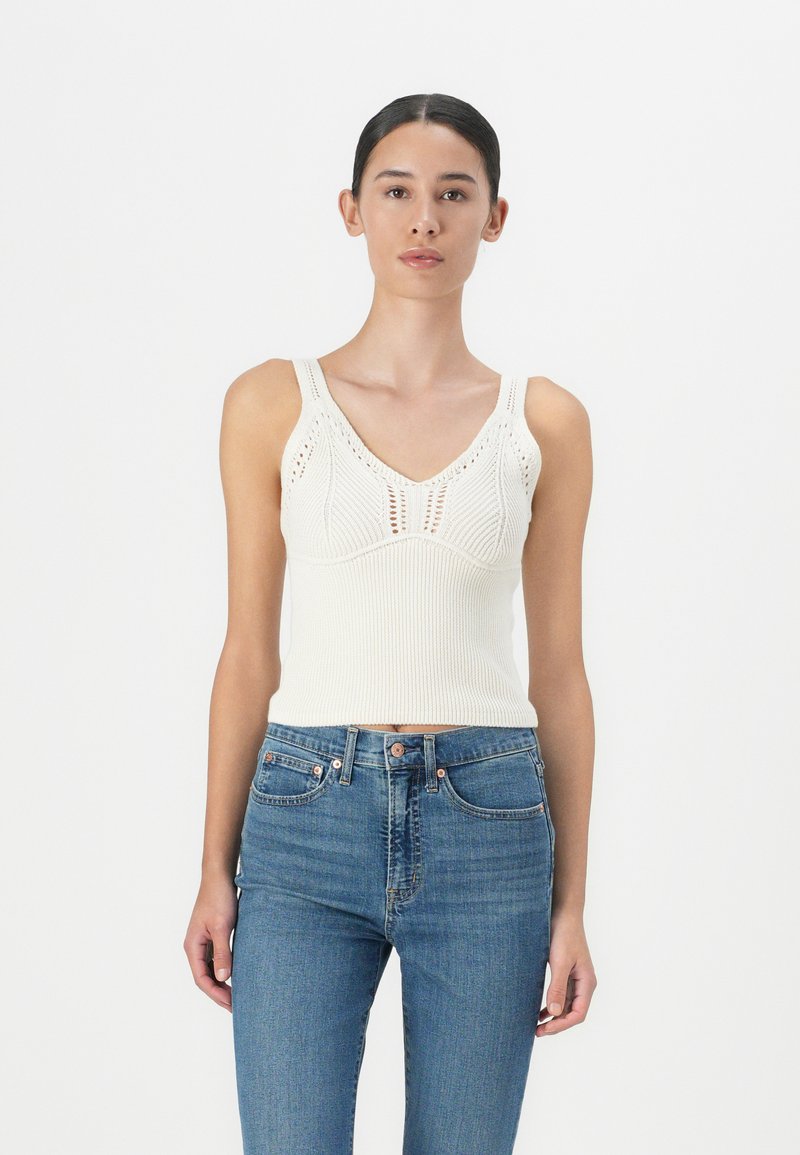 Hvit strikket tank topp med V-hals, ribbet tekstur og dekorativ åpenmønster. Kombinert med blå denim jeans.