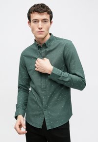 Grünes Hemd mit Knöpfen und Kragen, mit kleinen blauen Pünktchen und weicher Textur, kombiniert mit einer schwarzen Hose.