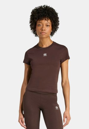 Donna con capelli ricci che indossa una maglietta aderente Adidas marrone scuro e pantaloni abbinati con piccoli loghi bianchi Adidas sul petto e sulla coscia.