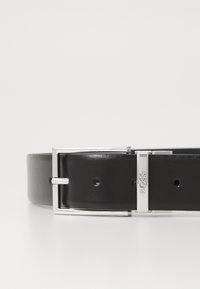 Ceinture en cuir noir avec une texture lisse, boucle rectangulaire en argent et insigne de la marque. Dispose de plusieurs trous pour des ajustements de taille.