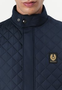 Marineblå quiltet jakke med høy krage, trykknapplukking og et gulllogo-patch på venstre side. Den har en glatt, teksturert overflate.