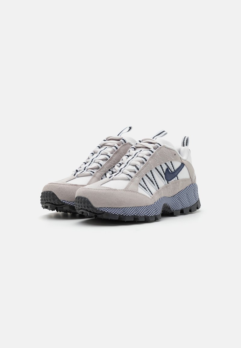 Les chaussures de sport présentent un mélange de daim gris et de maille blanche, avec une semelle en caoutchouc noir texturé et des accents distinctifs en bleu marine.