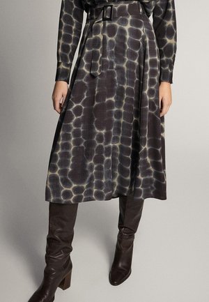 Femme portant une jupe midi noire et grise à motifs avec une ceinture assortie et des bottes en cuir marron foncé montantes jusqu'aux genoux.
