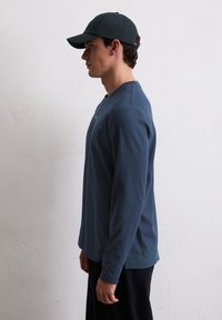 Ναυτικό μπλουζάκι με μακριά μανίκια από μαλακό ύφασμα με στρογγυλή λαιμόκοψη. Συνδυασμένο με ένα σκούρο καπέλο, το άτομο στέκεται μπροστά σε έναν ανοιχτό τοίχο με υφή.