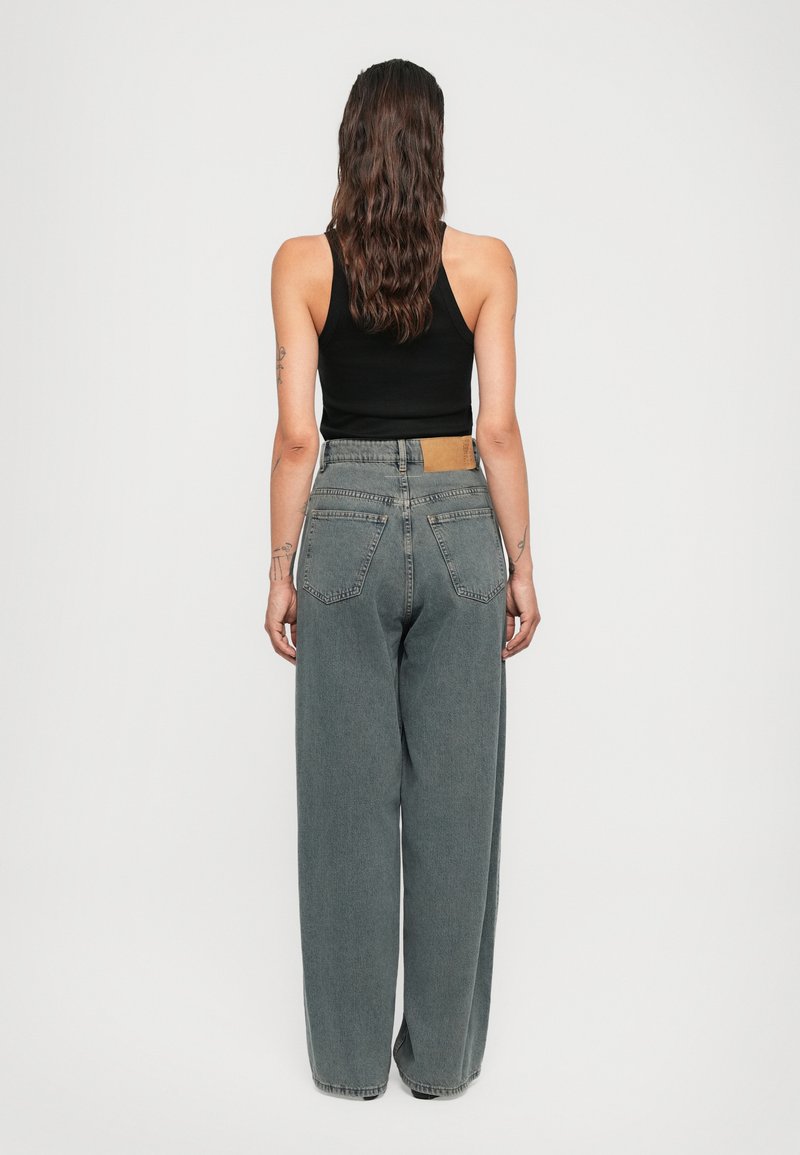 Kvinna med långt vågigt hår iförd en svart ärmlös topp och högmidjade grå jeans med vida ben, stående mot en enfärgad bakgrund.