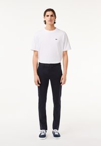 Camiseta de algodón blanca con un pequeño logo verde, combinada con pantalones slim-fit azul marino y zapatillas azul marino con cordones blancos, de pie frente a un fondo claro.