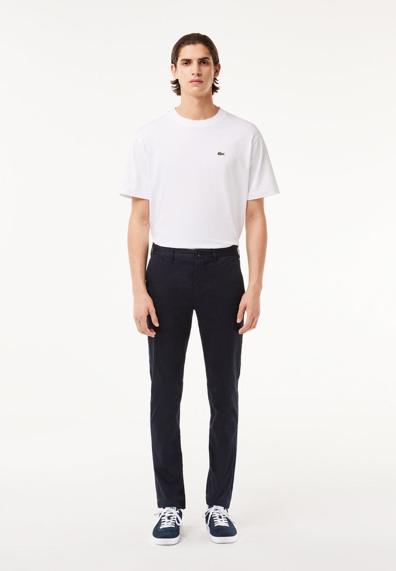 Camiseta de algodón blanca con un pequeño logo verde, combinada con pantalones slim-fit azul marino y zapatillas azul marino con cordones blancos, de pie frente a un fondo claro.