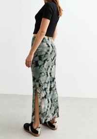 Frau in einem schwarzen kurz geschnittenen T-Shirt, einem grün-schwarzen Batik-Maxikleid mit seitlichem Schlitz und schwarzen und weißen Sneakers, die auf einem grauen Boden steht.