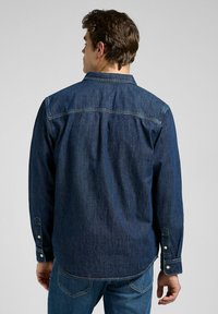 Camisa de mezclilla abotonada en azul oscuro, con mangas largas y un detalle de yugo. Ajuste estructurado y casual con un dobladillo ligeramente curvado.