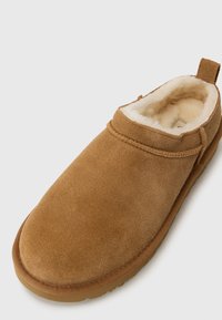 Ruskeat mokkasarja slippersit, joissa on pehmeä kermaisen värinen turkisvuori. Hialesin muotoilu ja joustava kumipohja, jossa on takana lenkki helpompaa pukemista varten.