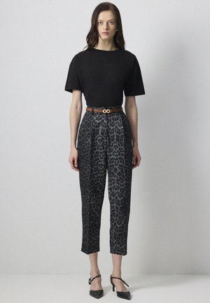 Naine kannab musta lühikeste varrukatega topi, halli kõrge vöökohaga leopardimustriga pükse pruuni vööga ning musti teravate ninadega kontsakingi, seisab ühevärvilise tausta ees.