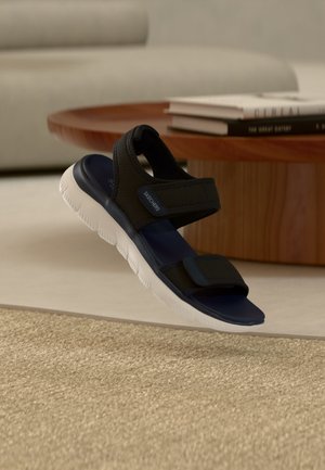 Czarna i granatowa sandałka Skechers z regulowanymi paskami i białą elastyczną podeszwą, umieszczona obok drewnianego stołu z poukładanymi książkami.