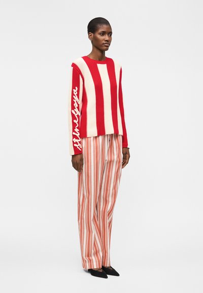 Γυναίκα που στέκεται, φορώντας πουλόβερ με κόκκινες και λευκές ρίγες και κείμενο στο μανίκι, συνδυασμένο με παντελόνι καμπάνα σε πορτοκαλί και λευκές ρίγες και μαύρες γόβες.