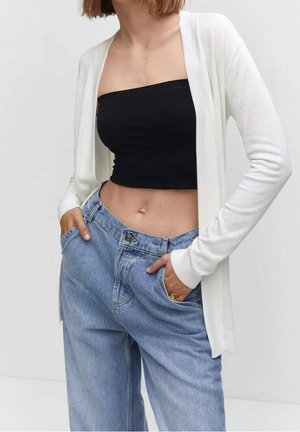 Femme portant un top bandeau noir, un cardigan blanc à manches longues et un jean bleu ample, les mains dans les poches, sur un fond uni.