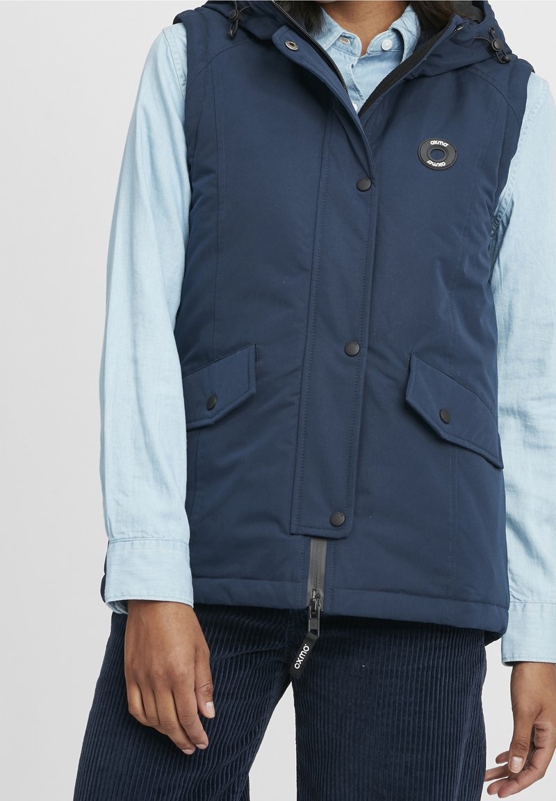 Marinblå väst med huva, tryckknappar, två framfickor och dragkedjestängning. Tillverkad av ett slätt, hållbart tyg.