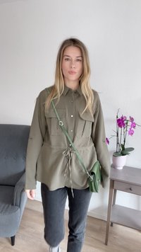 Olivegrünes Langarmhemd mit zwei Vordertaschen, Kordelzug in der Taille, kombiniert mit dunklen Hosen und einer kleinen grünen Umhängetasche.
