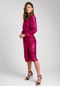 Fuksjowa sukienka zapinana na cekiny z długimi rękawami, paskiem w talii, długością do kolan i delikatnym połyskiem, zestawiona z czarnymi sandałami na wysokim obcasie.