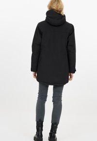 Sort hætteparka med en længere skæring, glat stof og justerbare manchetter. Bæres over grå slim-fit jeans og sorte ankelstøvler.