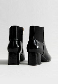 Botines negros hasta el tobillo con un diseño elegante, material brillante de charol, tacón ancho y paneles de tela en contraste, con cierre de cremallera en la parte posterior.