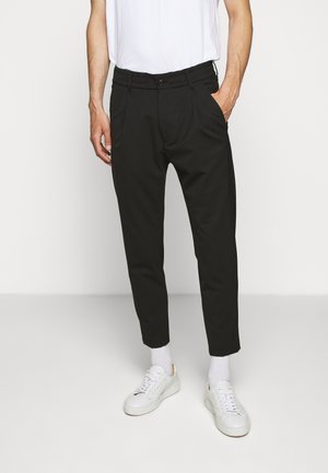 Mann trägt schwarze maßgeschneiderte Hose, weiße Turnschuhe, weiße Socken und ein weißes Hemd und steht mit einer Hand in der Tasche vor weißem Hintergrund.