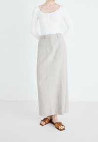 Person trägt langen beigefarbenen Rock, weißes langärmliges Oberteil und braune Sandalen, steht vor schlichtem weißem Hintergrund.