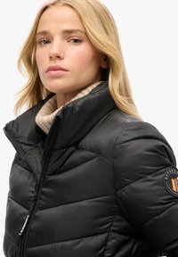 Czarna kurtka puchowa z wysokim kołnierzem i ukośnym przeszyciem. Posiada logo na rękawie oraz gładką, błyszczącą fakturę.