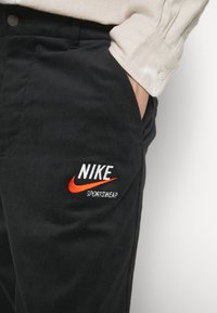 Black corduroy pants with a prominent white and orange "Nike Sportswear" embroidered logo on the pocket, featuring a textured fabric.