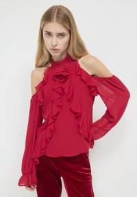 Rotes Chiffon-Oberteil mit Rüschen-Details, hohem Kragen und kalten Schultern. Lange Ärmel, weiche Textur, kombiniert mit roten Samthosen.