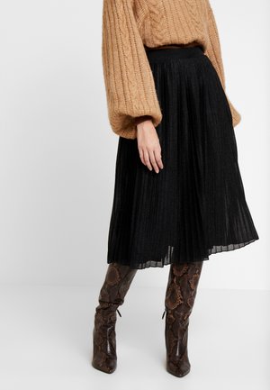 Kobieta ubrana w beżowy sweter z grubym warkoczowym splotem, czarną plisowaną spódnicę midi i brązowe kozaki za kolano w wężowy wzór, stojąca na białym tle.