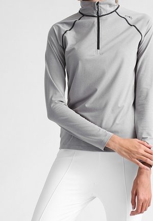 Persona che indossa un pullover grigio a maniche lunghe con zip frontale e pantaloni bianchi aderenti, in piedi con una mano che tocca il braccio opposto.