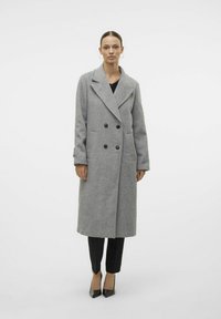 Cappotto doppiopetto in misto lana grigia con colletto a punte, bottoni neri, tasche laterali e lunghezza al ginocchio. Indossato con pantaloni neri.