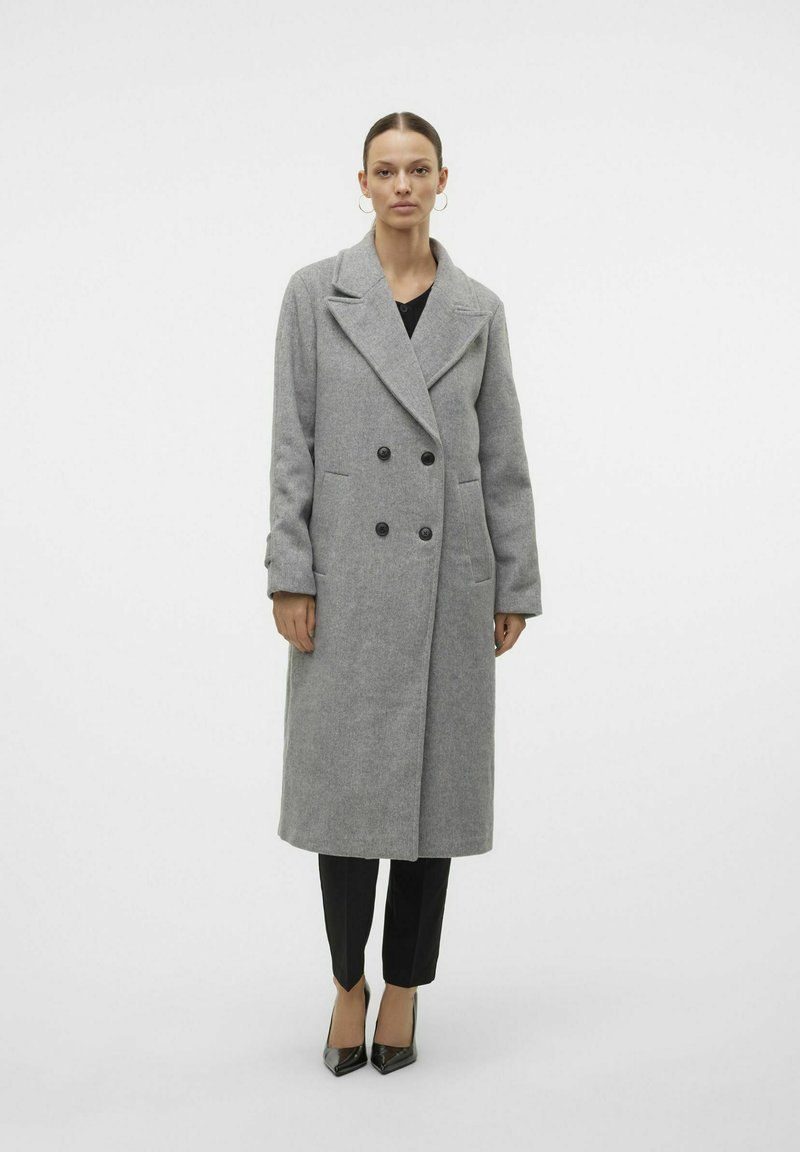 Cappotto doppiopetto in misto lana grigia con colletto a punte, bottoni neri, tasche laterali e lunghezza al ginocchio. Indossato con pantaloni neri.