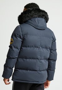 Jūrų mėlynos pūkinės striukės su dirbtiniu kailiu aptrauktomis gaubtu, siuvinėtu dizainu ir pleistru ant rankovės. Sklandi tekstūra ir šilumos izoliacija.