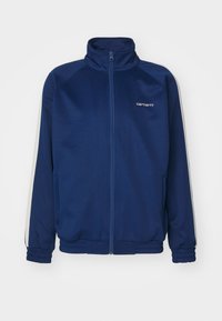 Tummansininen vetoketjullinen urheilujakku, jossa on korkea kaulus, joustavat hihansuut, valkoiset sivu raidat ja pieni valkoinen logo vasemmassa rinnassa.