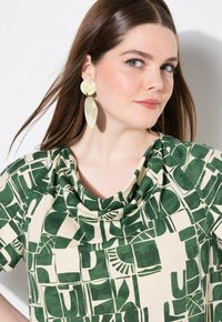 Blusa de manga corta con un patrón abstracto verde sobre un fondo beige claro, con un escote drapeado y una textura de tela suave.