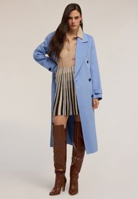 Cappotto di lana azzurro chiaro con bottoni doppi, camicia beige e gonna a righe bianco e nera abbinata a stivali marroni al ginocchio.