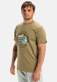 Olivově zelené bavlněné tričko s kruhovým grafickým motivem vln, hor a západu slunce se textem "Quiksilver Snowboarding Co."