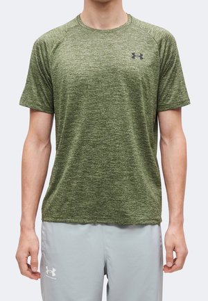 Mann trägt grünes meliertes kurzärmeliges Sportshirt sowie hellgraue Under Armour-Shorts, steht vor einfarbigem Hintergrund.