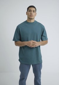 Turkizna bombažna majica s sproščenim krojem, kratkimi rokavi, okroglo ovratnikom in gladko teksturo, v kombinaciji z modrimi jeans hlačami.