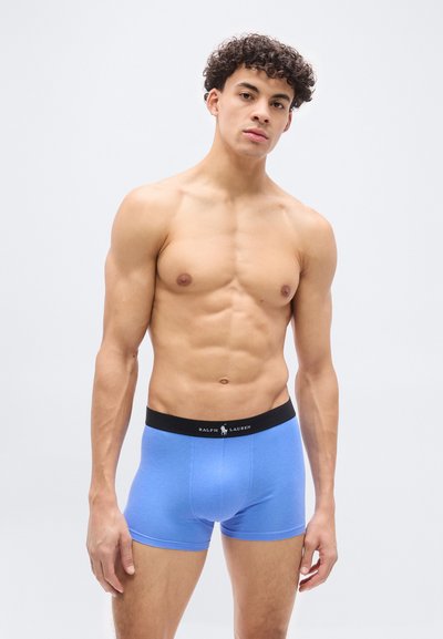 Νέος άνδρας με σγουρά μαλλιά φοράει μπλε ανδρικά εσώρουχα boxer με μαύρη ζώνη Ralph Lauren, στέκοντας μπροστά σε ένα απλό φόντο ανοιχτού χρώματος.