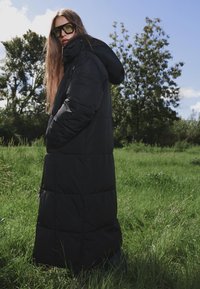 Dugi crni puffer kaput s kapuljačom, s horizontalnim šavovima, dva bočna džepa i detaljem sa zatvaračem. Lagan materijal, pogodan za hladno vreme.